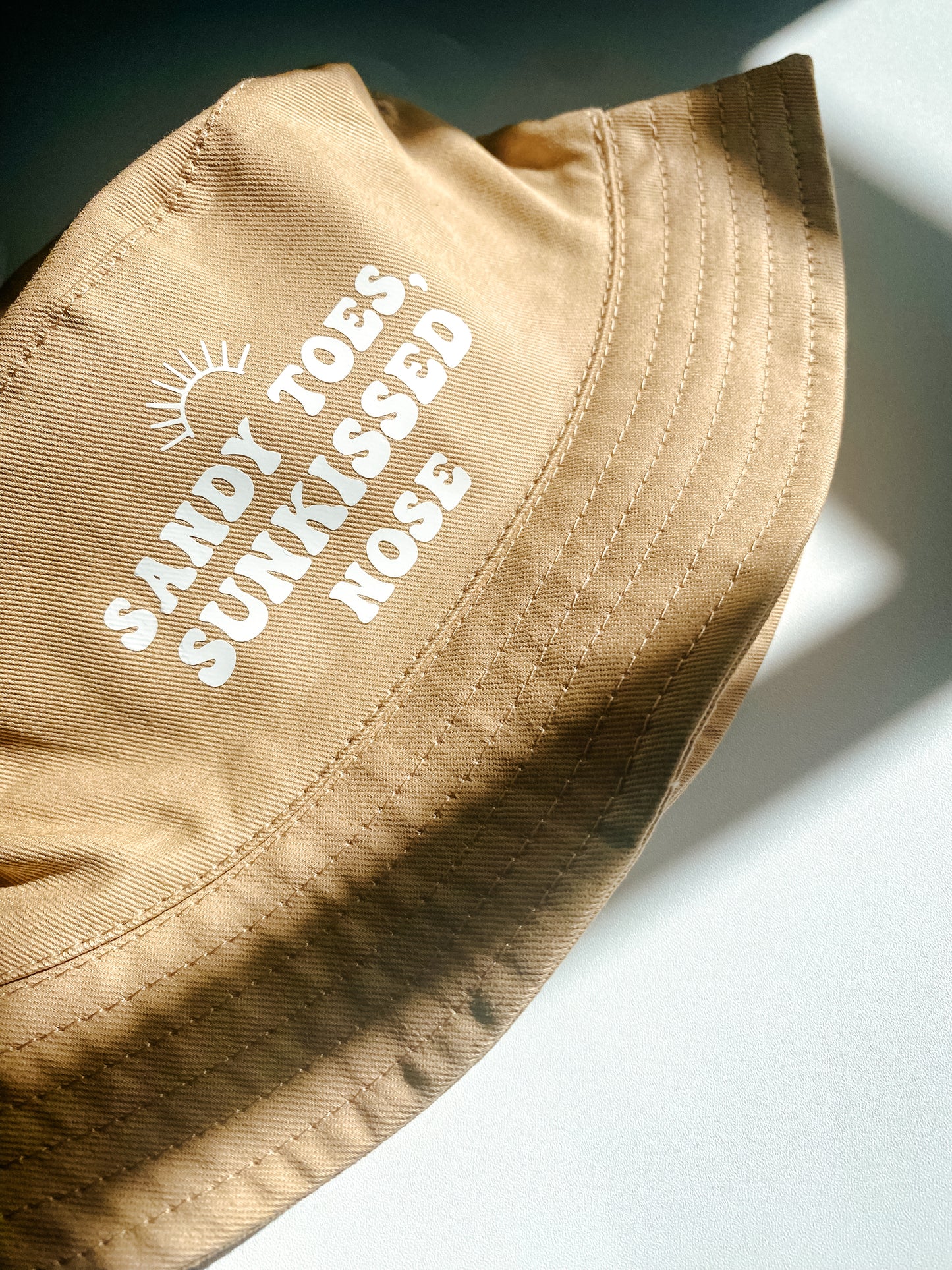 Beige summer bucket hat for baby with ‘sandy toes, sunkissed nose’ text printed slogan and small rainbow above. Text in white bubble font. 