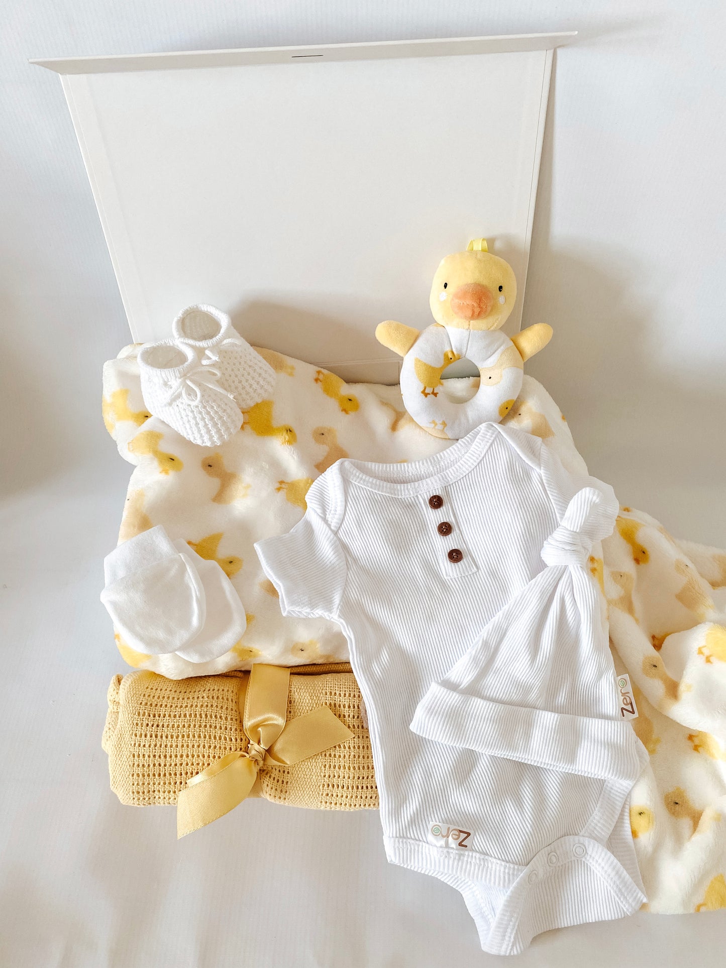 Baby gift box with items white baby hat, white baby vest, white pair of mittens, white knit booties, yellow cellular blanket, duck ring rattle with duck print & duck print fleece throw.