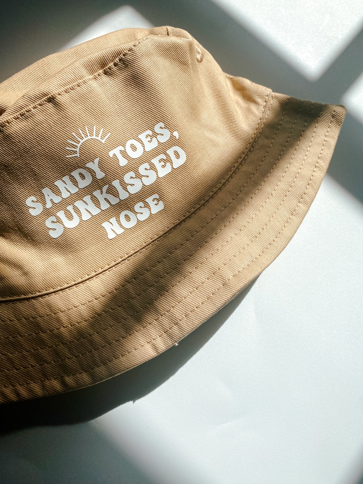 Beige summer bucket hat for baby with ‘sandy toes, sunkissed nose’ text printed slogan and small rainbow above. Text in white bubble font. 