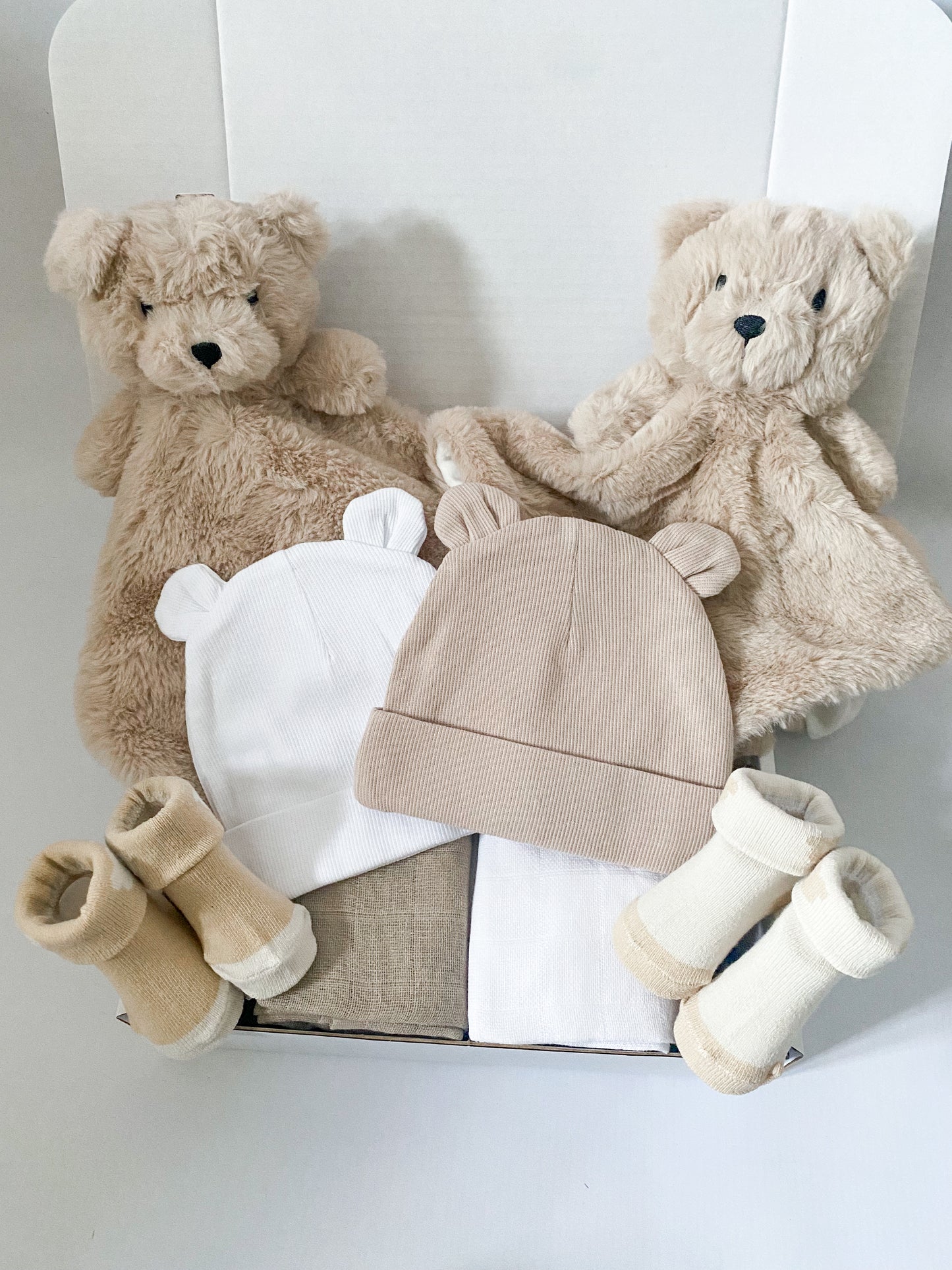 Baby twins gift box including two plush bear comforters, two bear ears beanie hats one in white one in beige, two pairs of alternate colour socks in beige and two square muslins one in white and one in beige.