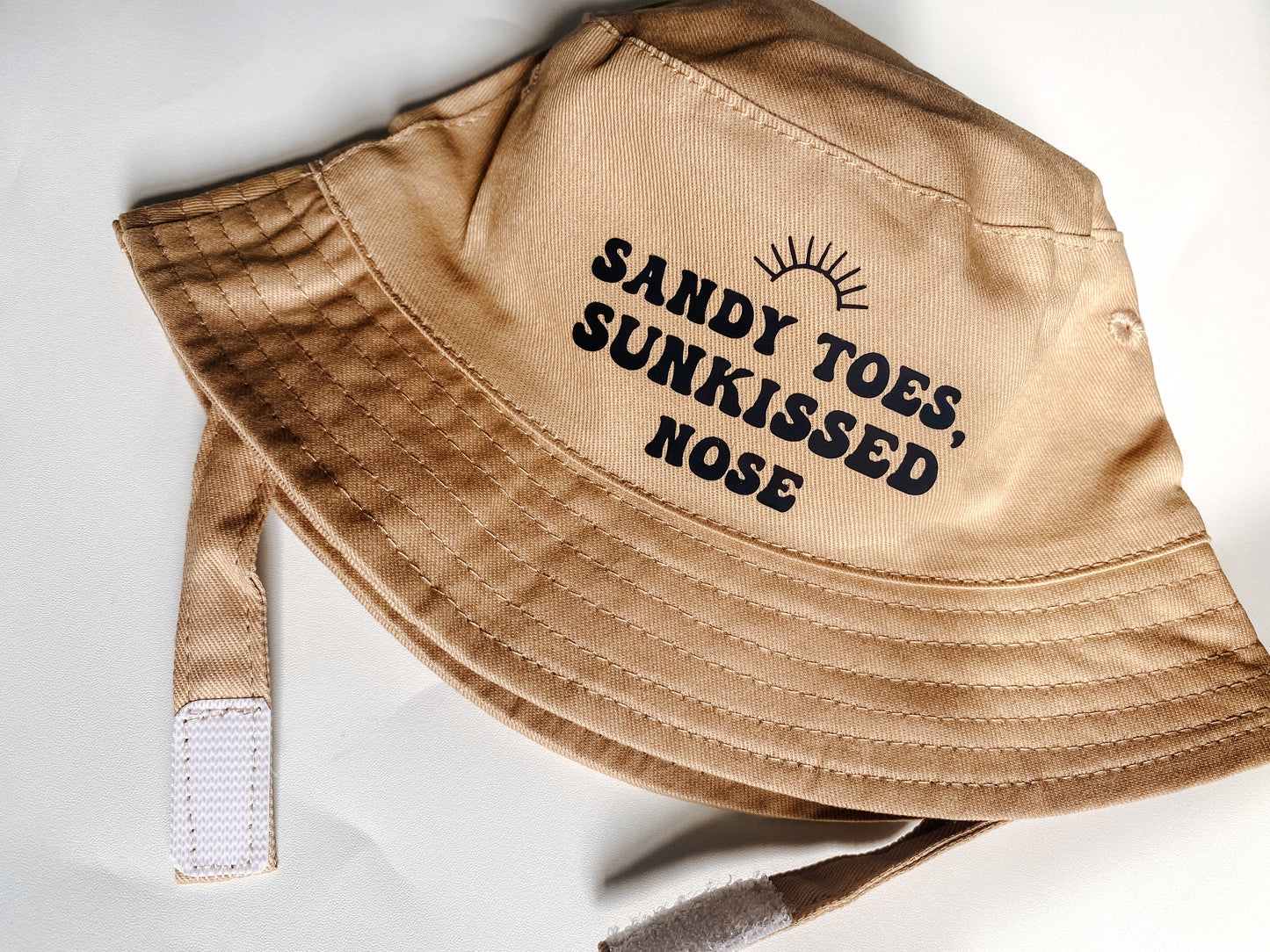 Beige summer bucket hat for baby with ‘sandy toes, sunkissed nose’ text printed slogan and small rainbow above. Text in black bubble font. 