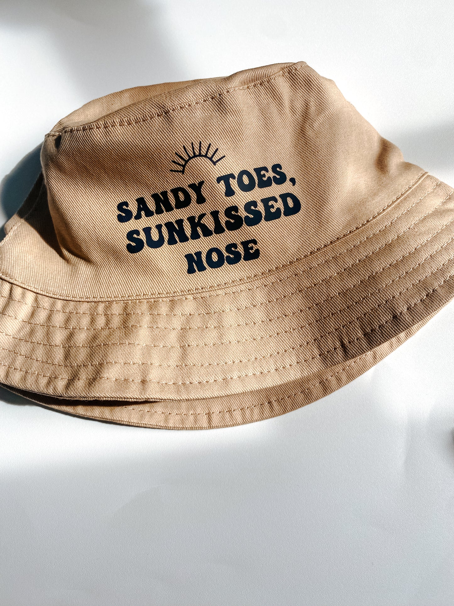 Beige summer bucket hat for baby with ‘sandy toes, sunkissed nose’ text printed slogan and small rainbow above. Text in black bubble font. 