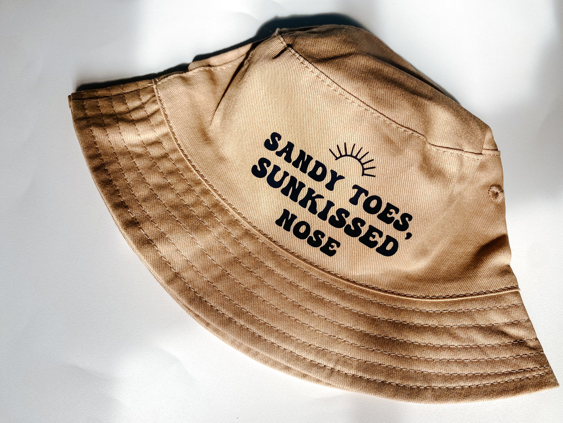 Beige summer bucket hat for baby with ‘sandy toes, sunkissed nose’ text printed slogan and small rainbow above. Text in black bubble font. 