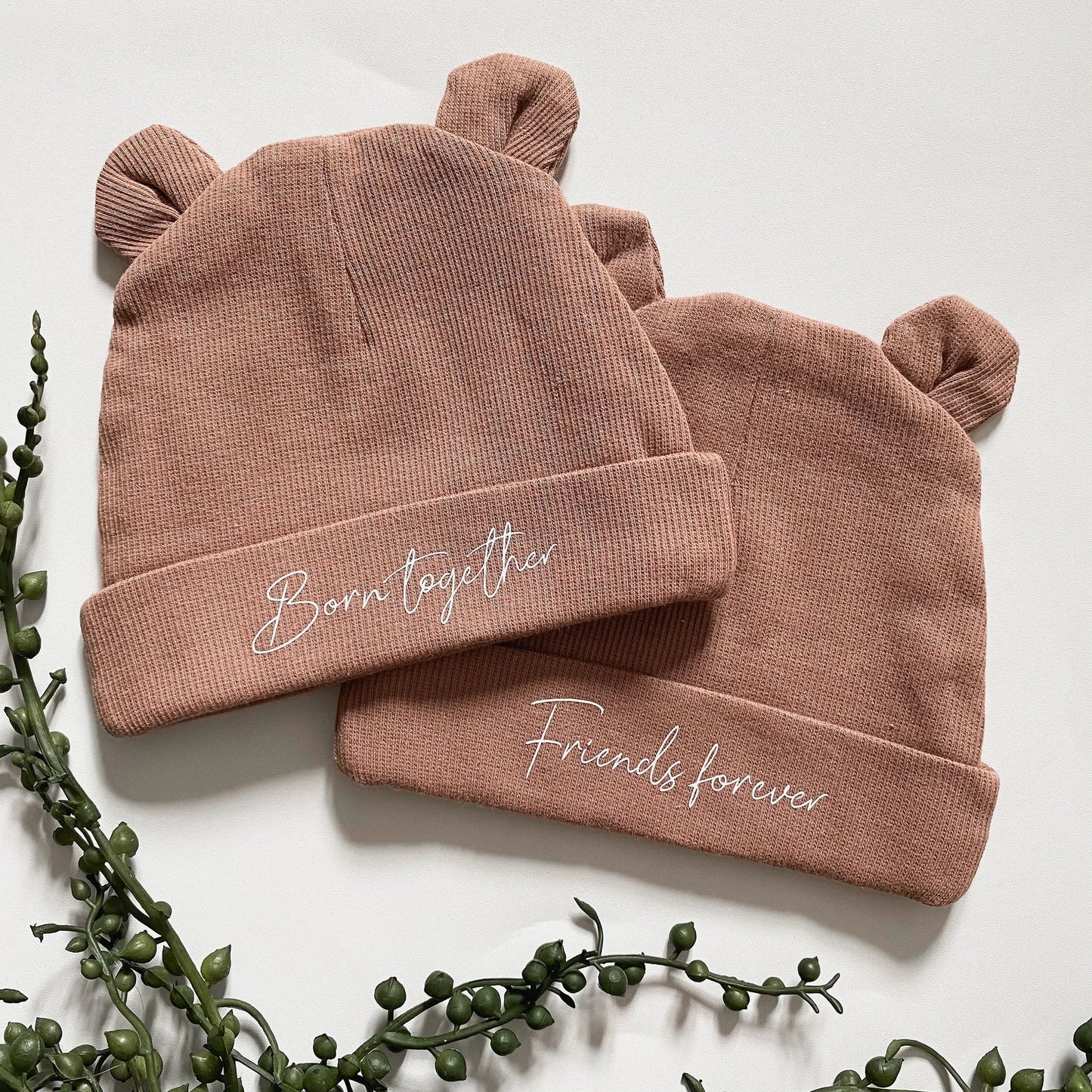 Brown baby bear ears hats one with the text “born together” and the other “friends forever”. White text italic font.