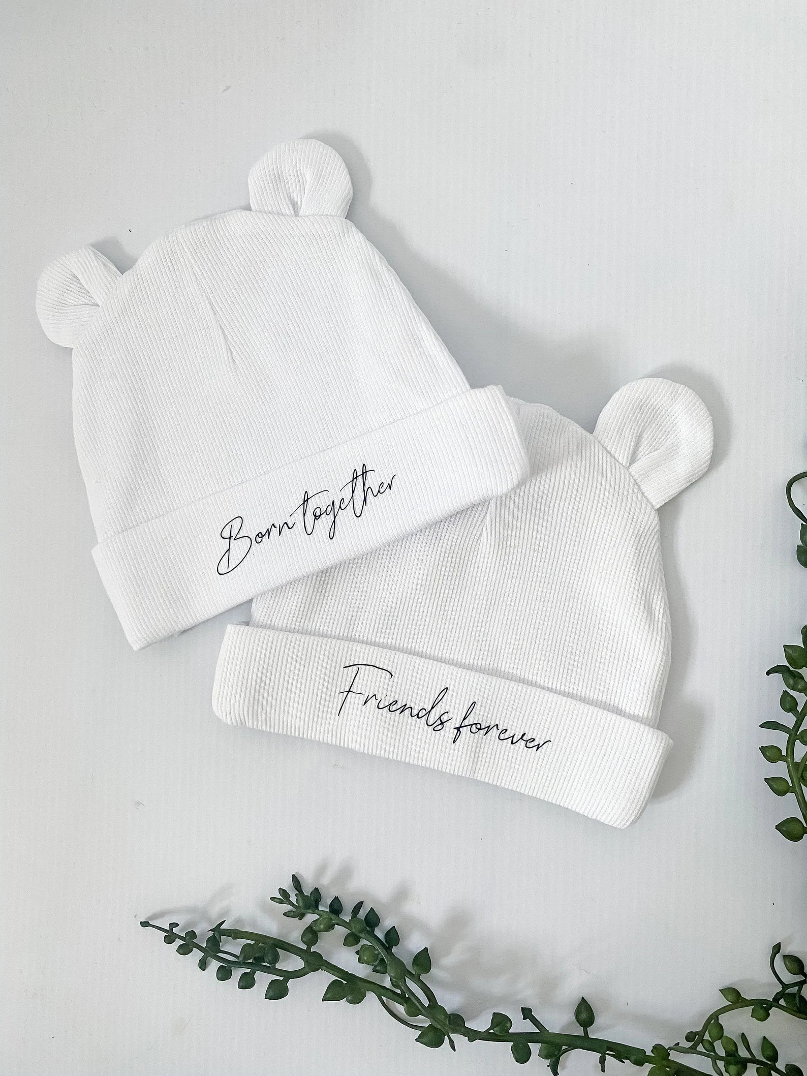 White baby bear ears hats one with the text “born together” and the other “friends forever”. Black text italic font.